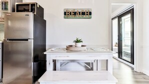 Una cocina con un refrigerador de acero inoxidable, una mesa blanca con una cesta y una planta, y un letrero que dice 'BEACH' en la pared.