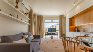 A modern living room with a grey sofa, wooden side tables, a glass dining table with wooden chairs, a woven rug, and a kitchen area with wooden cabinets and a built-in oven. - FLH Funchal Terrace Studio (Funchal)