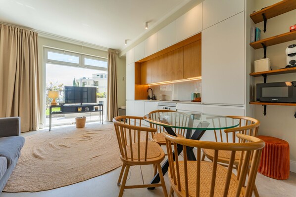 A modern living room with a glass table and wooden chairs, a sofa, and a large woven rug.