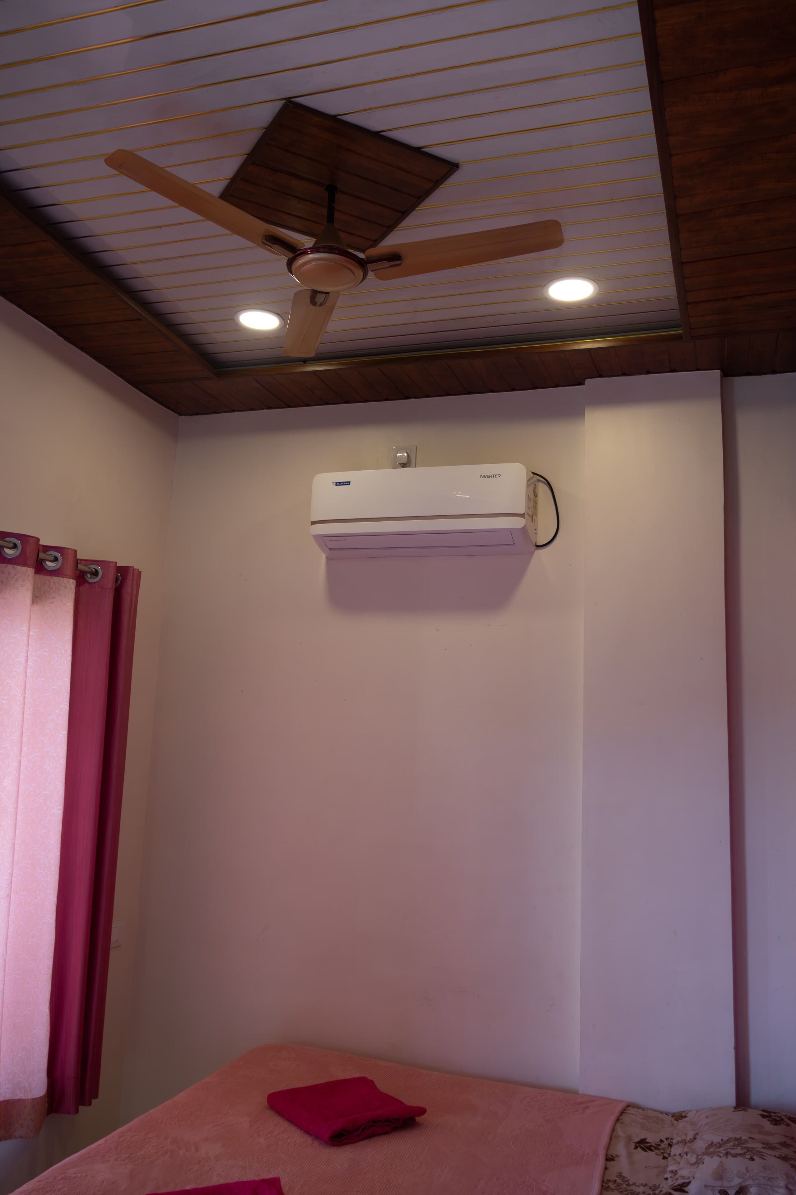 Una habitación con ventilador de techo, aire acondicionado, cama con manta rosa y ventana con cortinas.
