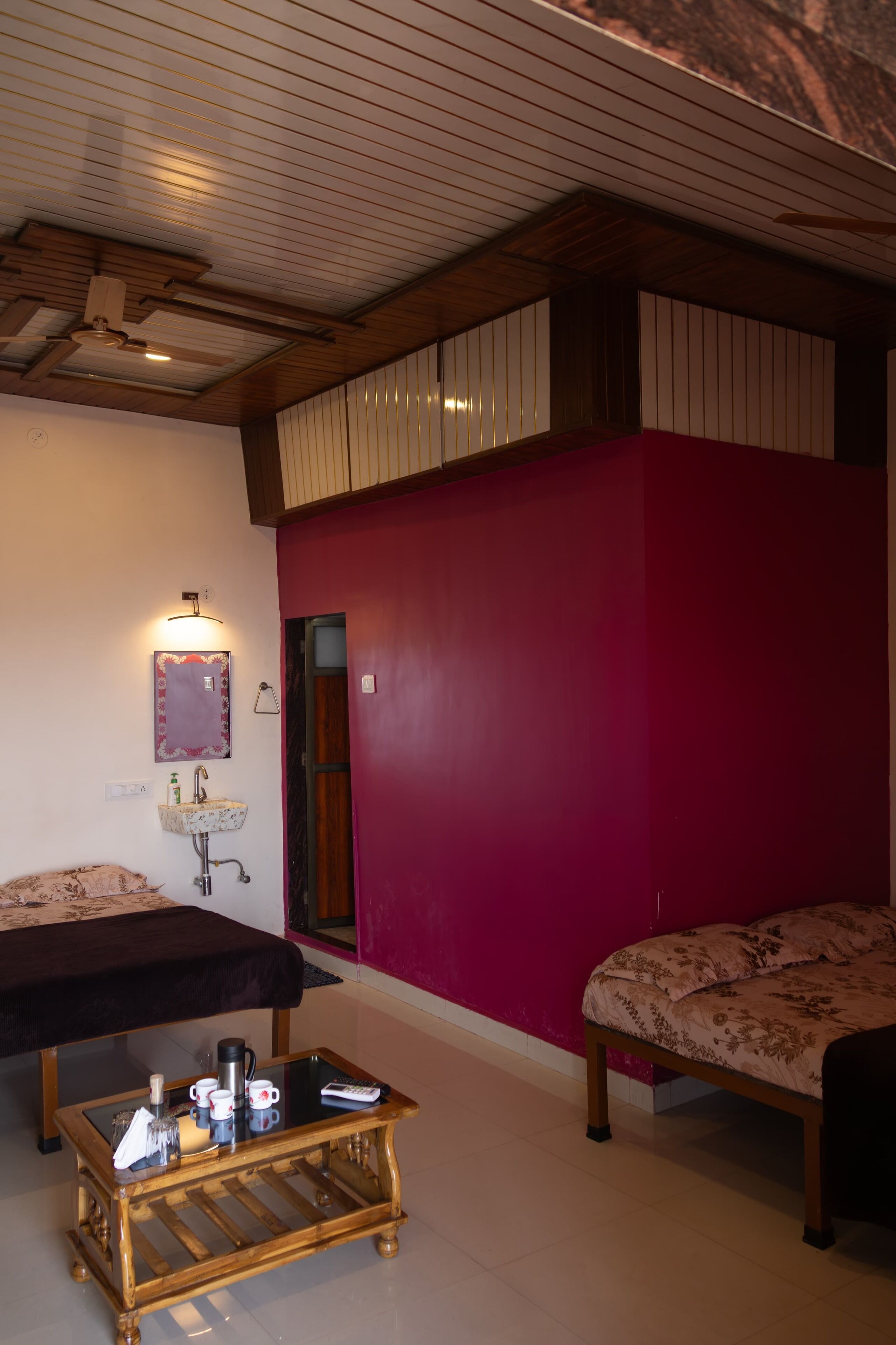 Een kamer met een houten plafond, een rode muur, een bed, een nachtkastje met een lamp en een klein raam.