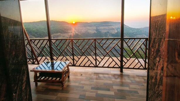A balcony with a wooden table and a view of the sunset over a hilly landscape. - Ovi cottage Matheran (Matheran)
