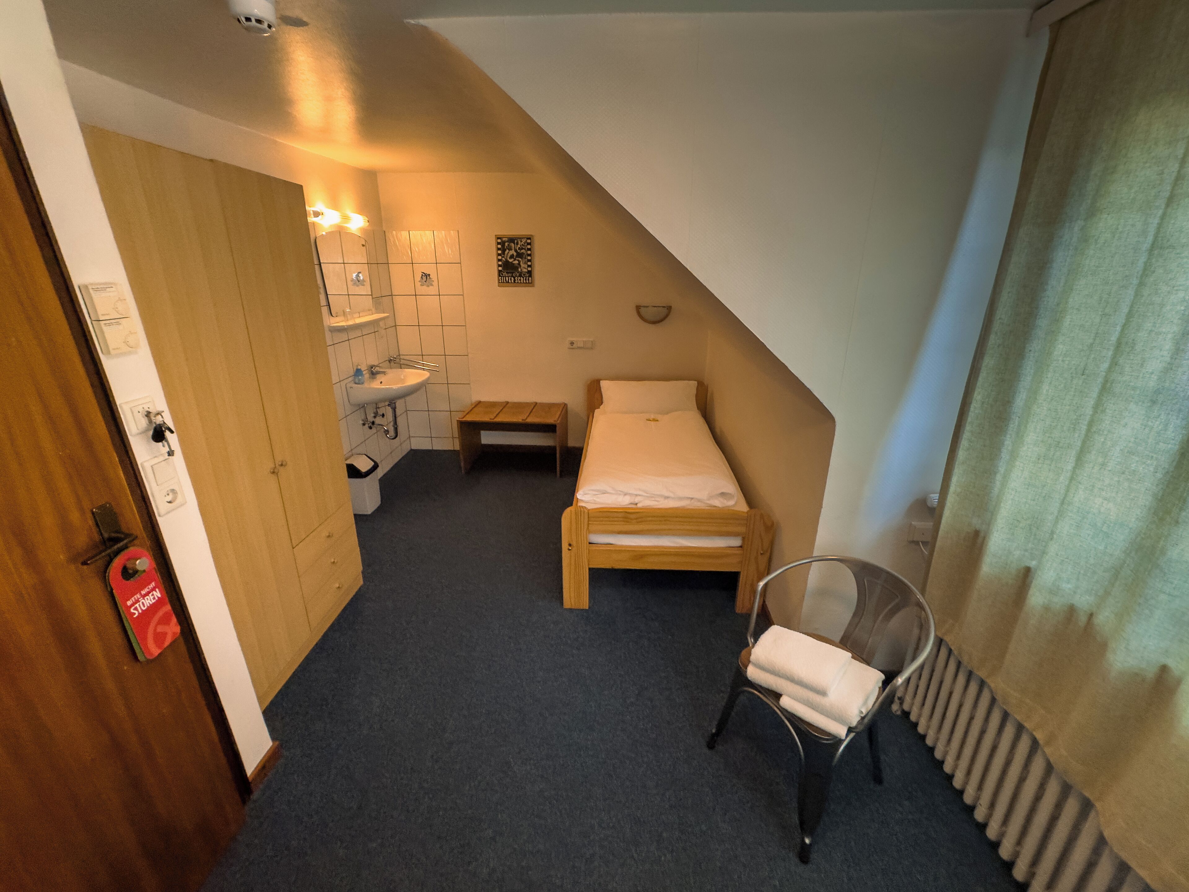Ein kleiner, schlichter Raum mit einem Einzelbett, einer hölzernen Bank, einem Stuhl, einem Heizkörper und einem Badezimmer mit Waschbecken und Spiegel.