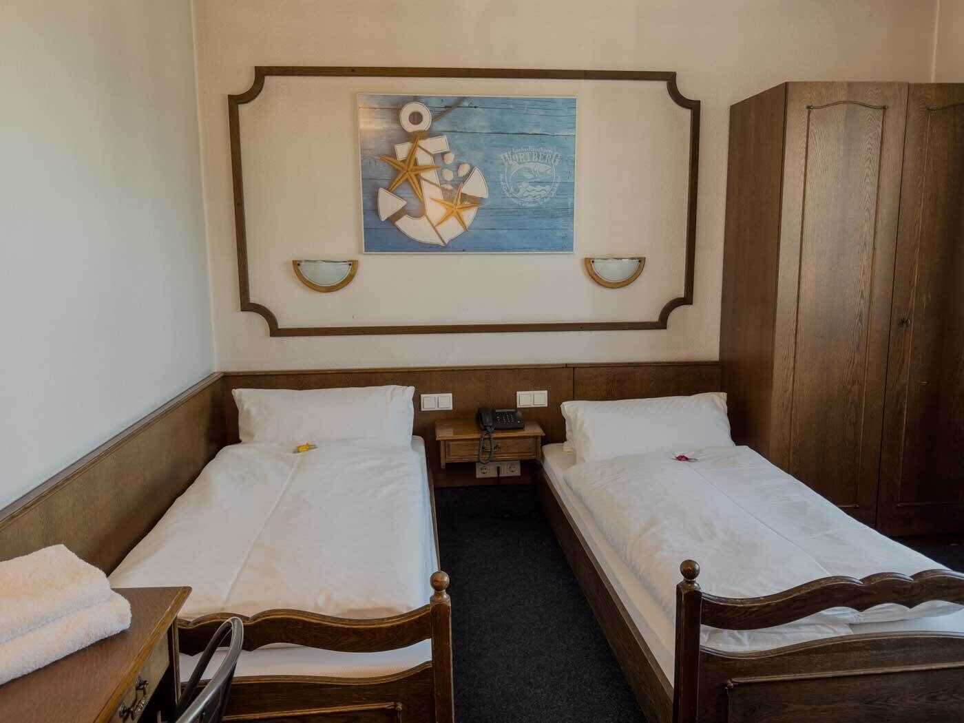 Una camera d'albergo con due letti singoli, un'armadio in legno, un comodino con una lampada e un'opera d'arte appesa al muro.