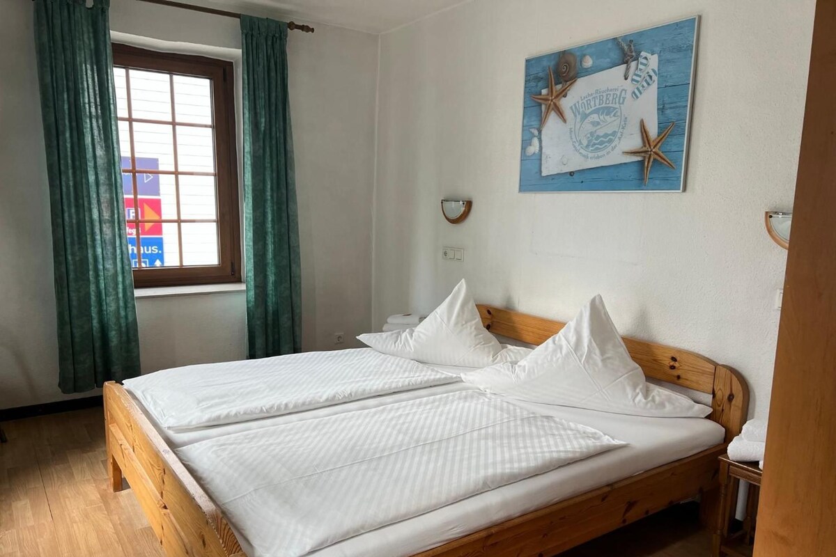 A double bed with white linens in a room with wooden flooring, a window with curtains, and a wall-mounted artwork.