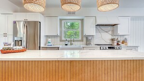 A modern kitchen with white cabinetry, a marble countertop, and a wooden island. There are wicker pendant lights, a stainless steel refrigerator, and a coffee maker.