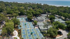 An aerial view of a coastal area with multiple tennis courts surrounded by dense greenery and a beach in the background.