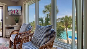 A room with two wicker chairs with striped cushions, a television on the wall, and a view of a pool and palm trees outside. - MyStay at 301 Shorewood - Oceanfront Views in S. Forest Beach + Beach Gear! (Hilton Head Island)