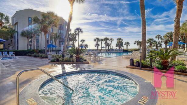 A pool area with lounge chairs, palm trees, and a building in the background. - MyStay at 301 Shorewood - Oceanfront Views in S. Forest Beach + Beach Gear! (Hilton Head Island)