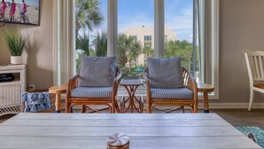 A room with two wicker chairs, a small table, a TV mounted on the wall, and a view of the beach and palm trees through large windows. - MyStay at 301 Shorewood - Oceanfront Views in S. Forest Beach + Beach Gear! (Hilton Head Island)