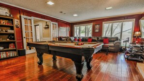 A pool table in a room with a red wall, a bookshelf, and a window with blinds. - Amazing 4,500 sq ft 2-story home in King's Gate. Perfect for large groups! (Lubbock)