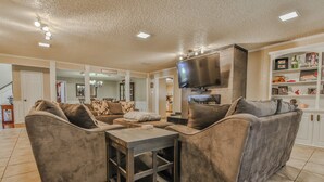 A spacious living room with a large flat-screen TV mounted on the wall, a comfortable seating area with sofas and a coffee table, and a kitchen area visible in the background. - Amazing 4,500 sq ft 2-story home in King's Gate. Perfect for large groups! (Lubbock)