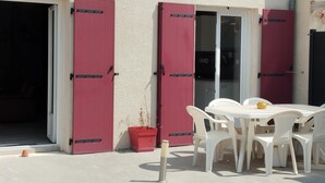Une terrasse avec une table et des chaises blanches, des volets rouges et un pot de fleurs.