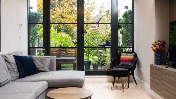 En moderne stue med en grå sofa, et rundt sofabord og en sort lænestol. Store vinduer giver udsigt over en have med forskellige planter og en træterrasse.