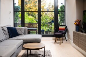 A modern living room with a grey sofa, a round coffee table, and a black armchair. Large windows offer a view of a garden with various plants and a wooden deck. - VESTO Cavendish Road (London)