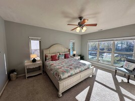 Un dormitorio con cama, mesita de noche, lámpara, ventilador de techo y ventana con vistas al exterior.