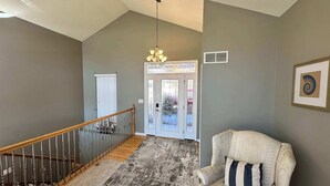 A staircase with a wooden handrail, a chandelier, a patterned rug, and a beige armchair with a striped pillow. - Lake It Easy - Waterfront Getaway With 7 Bedrooms! (Rocky Mount)