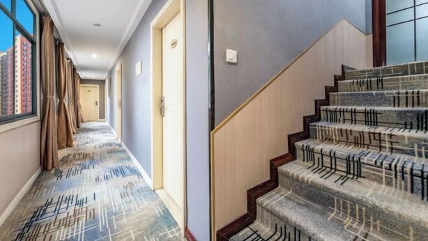 Un corridoio d'albergo con tappeto a motivi, una scala con gradini in legno e ringhiere in metallo, e una porta con interruttore della luce.