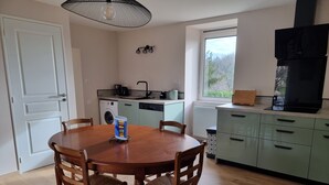 A kitchen with a dining table, chairs, a washing machine, a dishwasher, and a large window. - GÎTE SARAZAC (Mayac)