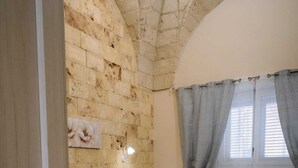 Un dormitorio con pared de piedra, cama con colcha estampada, cabecero gris, alfombra peluda negra y ventana con cortinas.