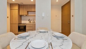 Un comedor con una mesa de mármol, platos blancos y cubiertos. Se ve una cocina al fondo con armarios de madera y una estufa.