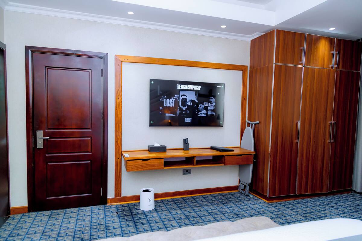 A hotel room with a wooden wardrobe, a television mounted on the wall, a wooden console table, and a patterned carpet.