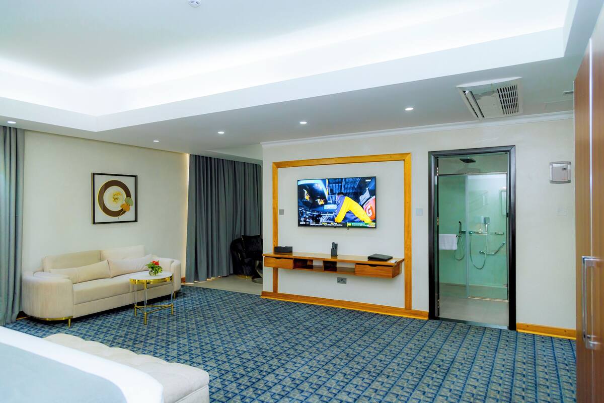A hotel room with a large flat-screen TV mounted on the wall, a sofa, a small coffee table, and a patterned carpet.