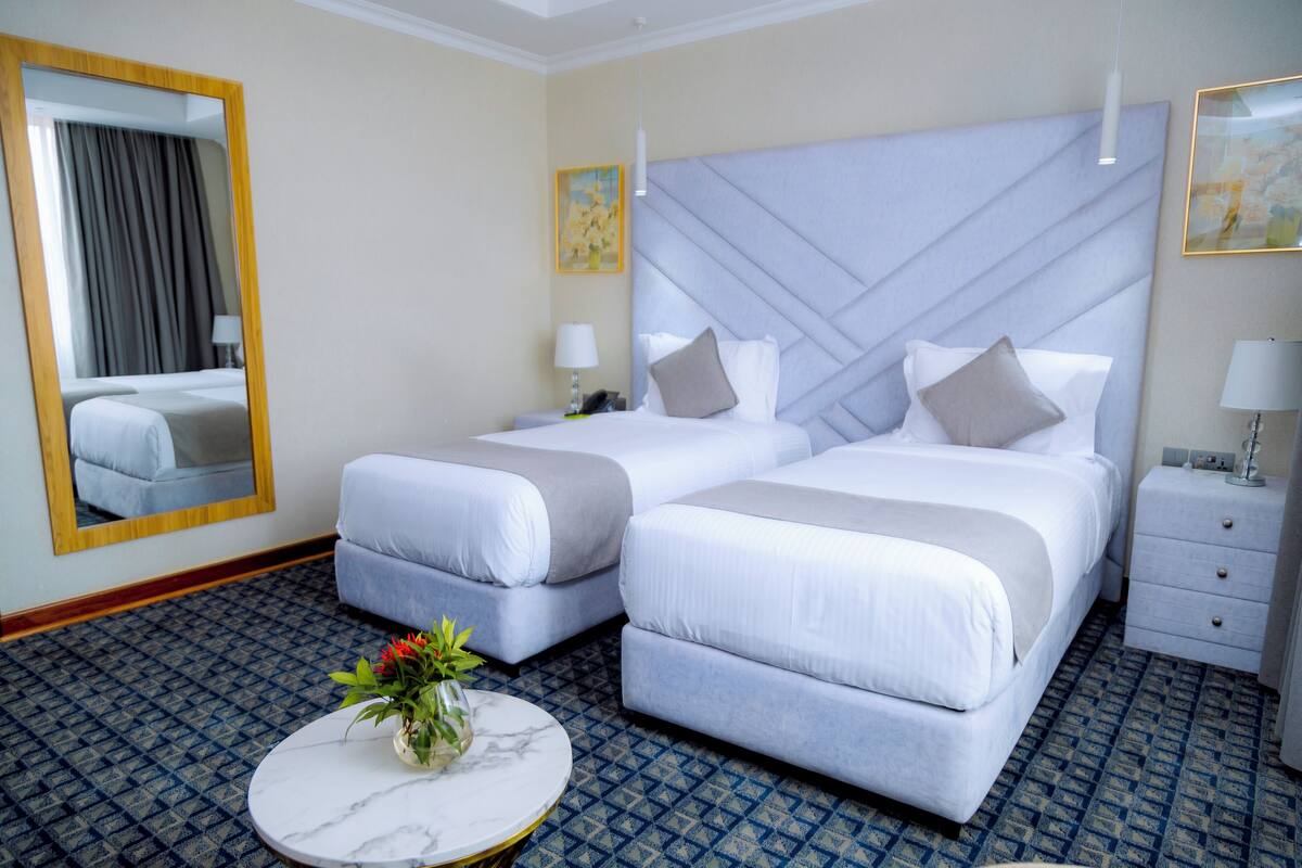 A hotel room with two beds, a round marble table with a vase of flowers, a large mirror, and a patterned carpet.