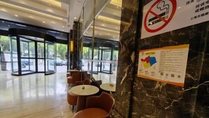 Un espace intérieur moderne avec des portes vitrées donnant sur l’extérieur, plusieurs tables rondes et des chaises orange, ainsi qu’un mur de marbre orné d’un panneau « interdiction de fumer » et de panneaux d’information.