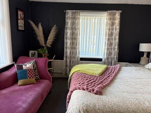 A hotel room with a bed, a pink sofa, a window with curtains, a radiator, and a lamp. - 5 Bed Villa - Sleeps 10 - Pets - Garden - Parking (Skegness)