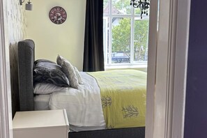 A bedroom with a bed, pillows, a bedside table, a wall clock, and a window with a view of trees and buildings.