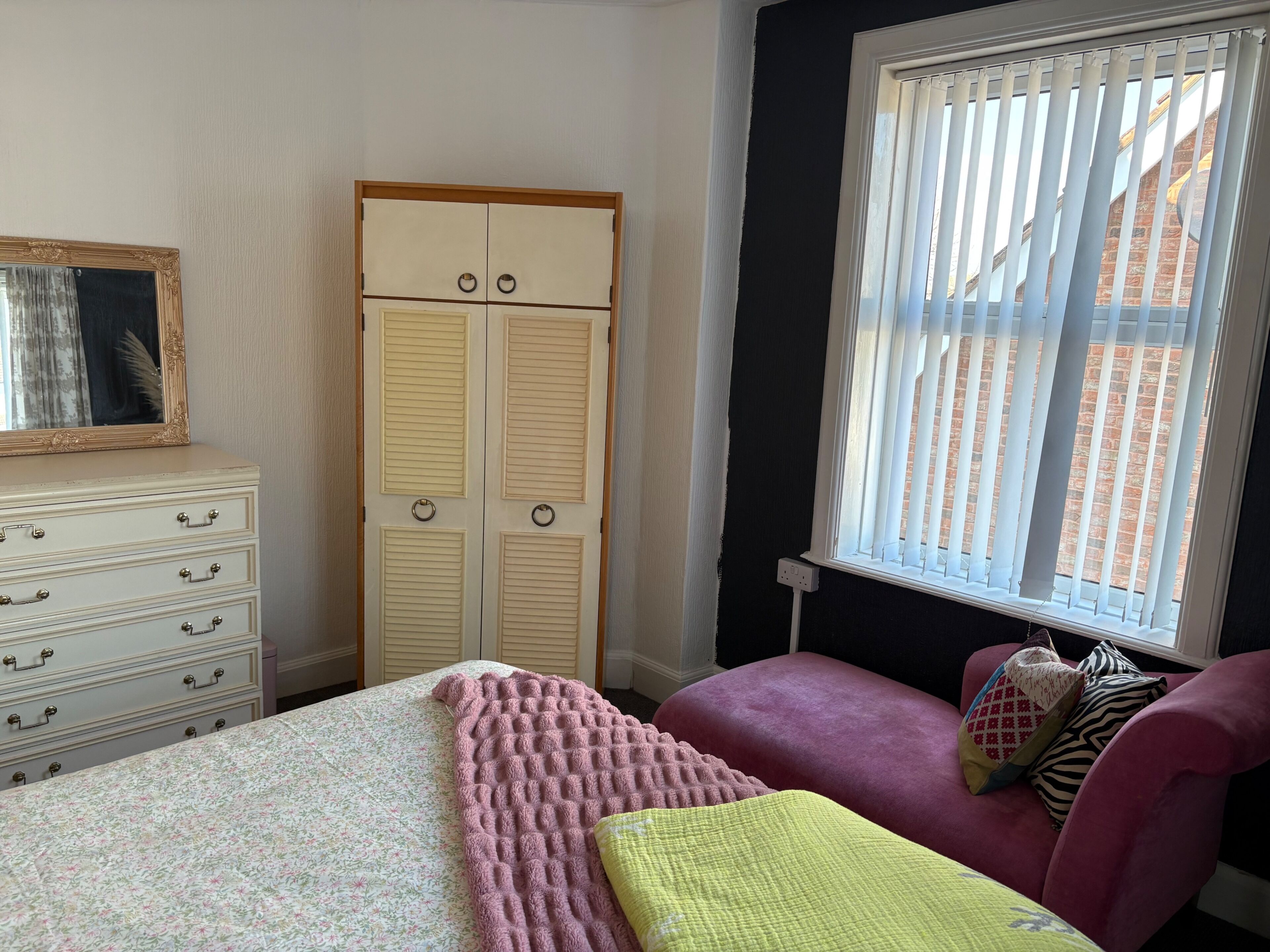 A bedroom with a bed, a dresser, a wardrobe, a window with blinds, and a chair with cushions.