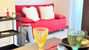 Un salon moderne avec un canapé rouge, des coussins blancs et moelleux, une table basse en verre et une bibliothèque contenant des livres et une télécommande.