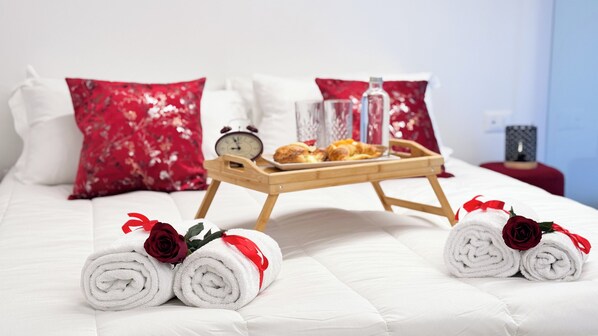 Una cama bien hecha con sábanas blancas, almohadillas rojas con estampado floral y una bandeja con pasteles y un vaso de agua.