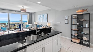 A modern kitchen with a large window overlooking a beach, a black countertop, and a built-in bookshelf. - Sterling Shores 616 (Destin)