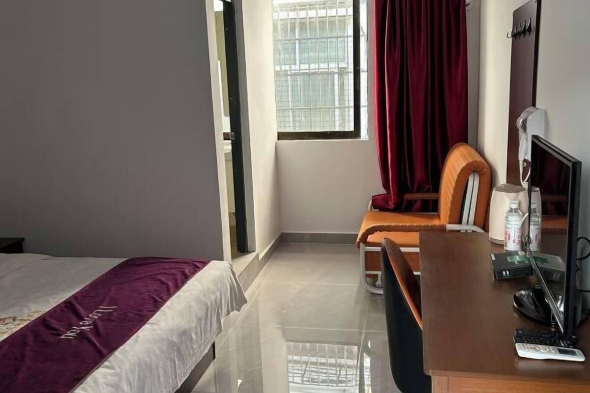 A hotel room with a bed, a desk with a computer, a chair, a window with curtains, and a bedside table with a lamp and a bottle.