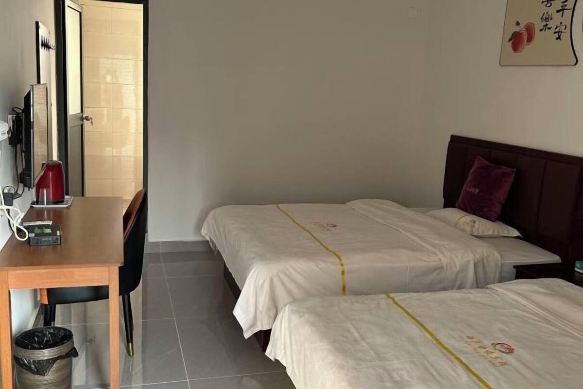 A hotel room with two beds, a wooden desk, a trash bin, and a wall-mounted TV.