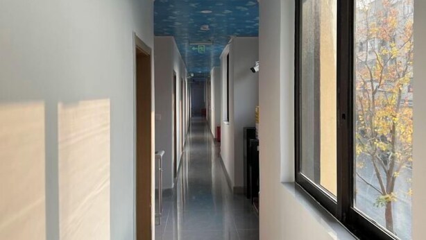 Un couloir avec un plafond bleu, de grandes fenêtres et une vue sur les arbres à l’extérieur.