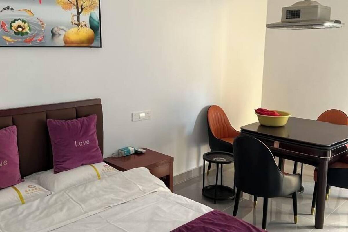 A hotel room with a bed, a dining table with chairs, a painting on the wall, and a small table with a bowl.