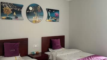 Quarto de hotel com duas camas, cada uma com almofada roxa e cobre branca. Três obras de arte na parede retratam cenas surreais.