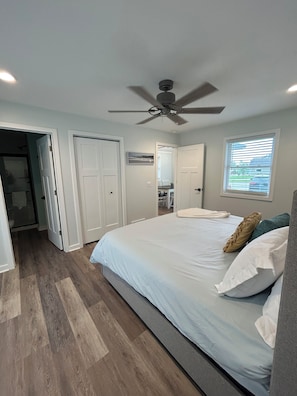 Um quarto com cama, ventoinha no teto, janela com persianas e porta de acesso a um banheiro.