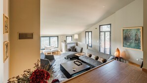 A spacious living room with a large sofa set, a coffee table, and a fireplace. There are multiple windows providing natural light and a view of the outdoors.