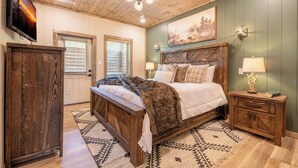 A bedroom with a wooden bed, a nightstand, a TV mounted on the wall, and a painting above the bed. - *Modern 3BD Cabin w/ Hot Tub + Arcade! Near TOWN!* (Pigeon Forge)