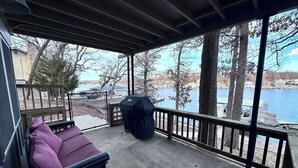 Un espace extérieur abrité, doté d’un banc rembourré, d’un barbecue et offrant une vue sur un lac et des bâtiments.