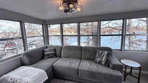 Un salon moderne avec un canapé d’angle gris, un plaid blanc et un tapis à motifs. On y trouve une petite table ronde et un luminaire au plafond original. De grandes fenêtres offrent une vue sur un plan d’eau et des arbres.