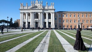 A large open square with a grand building featuring columns and statues in the background.