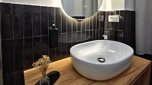 A modern bathroom with a large oval mirror, a white sink, and black tiled walls.