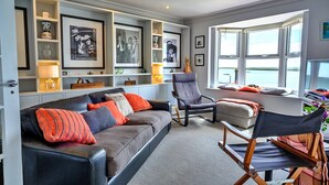 Een moderne woonkamer met een bank, fauteuils en een regisseursstoel. Er hangen ingelijste foto’s aan de muur, een raam met uitzicht en een plank met decoratieve objecten.