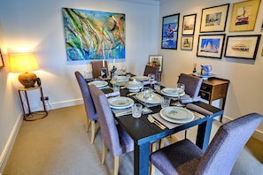 A dining room with a table set for six, surrounded by chairs, with artwork on the walls and a lamp on the side. - Apartment in Burnham With River Views (Burnham-on-Crouch)