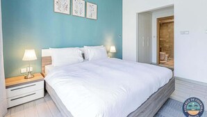Une chambre à coucher avec un grand lit, deux tables de chevet avec des lampes, une tête de lit en bois et une porte donnant sur une salle de bain.